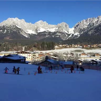 Eine verschneite Landschaft mit einer Skischule und mehreren Kindern im Skitraining. Im Hintergrund sind beeindruckende Berge und traditionelle Alpenhäuser zu sehen.
