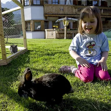 A cheerful girl is sitting in the garden, playing with a small black rabbit. In the background, a cozy wooden house can be seen.