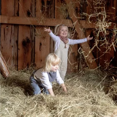 Two children are happily playing in a barn full of hay. The younger child is on the ground and the older child is standing upright and laughing.
