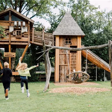 A wooden playground with a treehouse and a slide. Two children are happily running towards the play equipment.