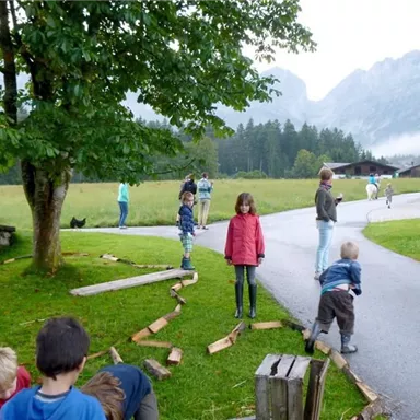 Eine Gruppe von Kindern spielt im Freien, umgeben von Bäumen und Bergen. Es ist eine ruhige und idyllische Szenerie auf einem Landhof.
