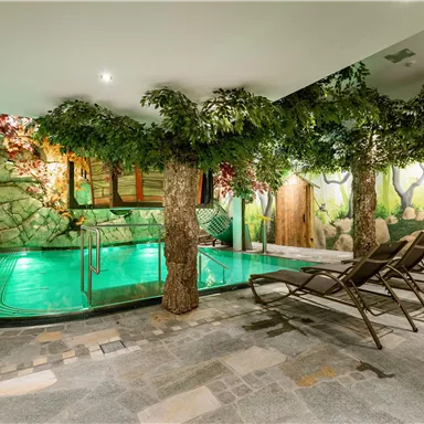 Ein entspannender Wellnessbereich mit einem grünen Pool und dekorativen Bäumen. Im Hintergrund sind gemütliche Liegen und eine natürliche, steinige Wand zu sehen.