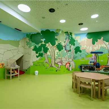 Ein farbenfroher Spielraum mit kindgerechten Wandmalereien und einem gemütlichen Fußboden. Es gibt Spieltische und eine Spielecke, ideal für Kinder.