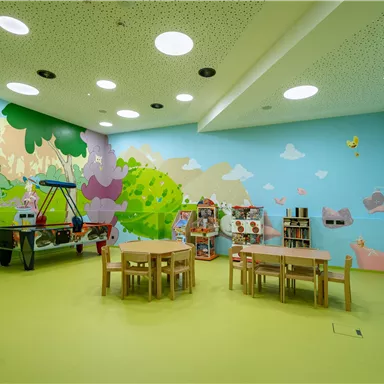 Ein bunter Spielraum mit freundlichen Wandmalereien. In der Mitte stehen Tische und Stühle zum Spielen und Lernen.