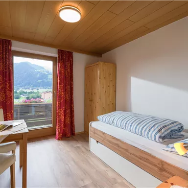 Ein einfaches Zimmer mit einem einzelnen Bett und einem Schreibtisch. Große Fenster bieten einen Blick auf die Berge.