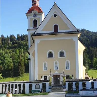 Eine malerische Kirche mit einem roten Turm und gelber Fassade. Umgeben von grünen Hügeln und Bäumen.