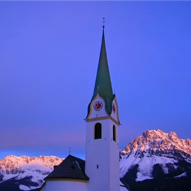 Eine Kirche mit einem hohen, spitzen Turm steht unter einem bunten Himmel. Im Hintergrund sind schneebedeckte Berge zu sehen.