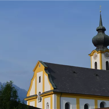 A pretty church with a yellow facade and a distinctive spire. In the background, there are mountains and a blue sky.