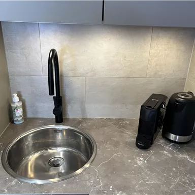 Eine moderne Küchenecke mit einem runden Edelstahlspülbecken und einem schwarzen Wasserhahn. Neben dem Spülbecken stehen eine Kaffeemaschine und ein Wasserkocher.
