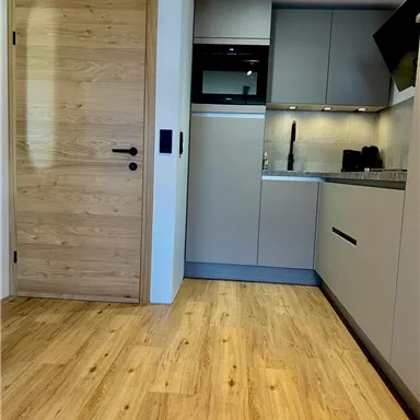 Eine moderne Küche mit hellen Möbeln und Holzfußboden. Die Wand ist teilweise mit grauen Schränken und einem Kochfeld ausgestattet.