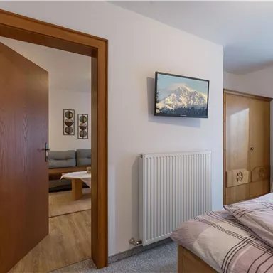 A cozy bedroom with a bed and a television on the wall. Through the open door, a living area can be seen.