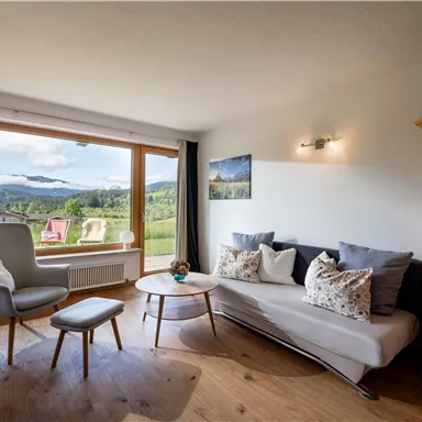 Ein modernes Wohnzimmer mit großen Fenstern und Blick auf eine grüne Landschaft. Es gibt gemütliche Möbel und viel Tageslicht.