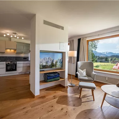 Ein modernes Wohnzimmer mit einer offenen Küche und großen Fenstern. Der Blick zeigt auf eine malerische Landschaft mit Bergen und strahlend blauem Himmel.