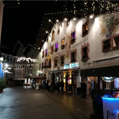 A picturesque street at night, illuminated by lights and adorned with festive decorations. The buildings are traditional and welcoming, with colorful windows.