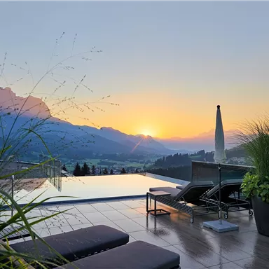 A beautiful pool with loungers and plants on a terrace. In the background, mountains and a breathtaking sunset can be seen.