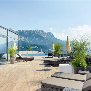 A modern terrace with loungers and sun umbrellas offers a stunning view of the mountains. The clear sky and the green landscape create a relaxing atmosphere.
