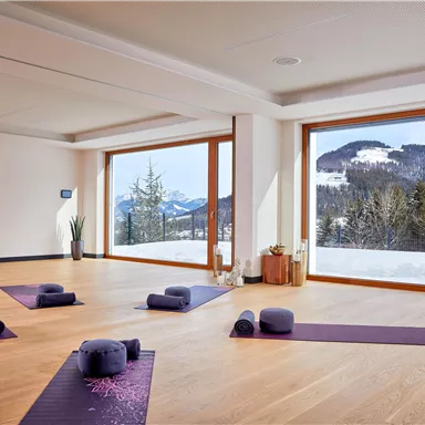 A bright yoga room with large windows overlooking snow-covered mountains. Yoga mats and cushions are spread across the floor, creating a serene atmosphere.