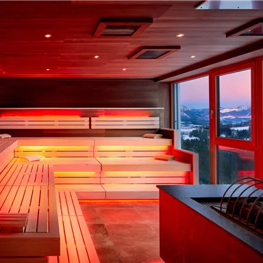 A modern sauna with soft red light. In the background, snow-capped mountains and a beautiful sunset can be seen.