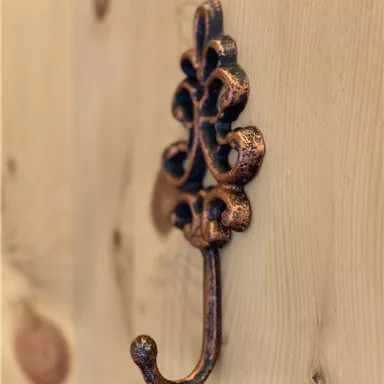 A decorative metal hook on a wooden wall. The hook has an artistic design and is in a copper color.