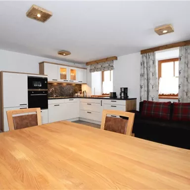 Eine moderne Küche mit einem großen Esstisch aus Holz. Der Raum ist hell und freundlich gestaltet, mit einem gemütlichen Sofa und Fenstern mit Vorhängen.