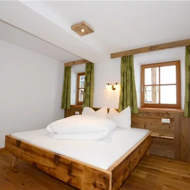 Ein modernes Schlafzimmer mit einem großen Holzbett und weißen Kissen. Die Wände sind hell, und die Fenster haben grüne Vorhänge.
