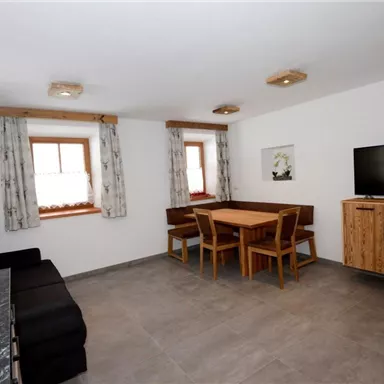 Ein modernes Wohnzimmer mit einem Sofa, einem Esstisch und Stühlen. An der Wand befindet sich ein Fernseher, und die Fenster sind mit Vorhängen dekoriert.