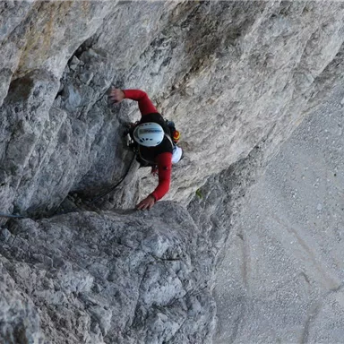 Ein Kletterer erklimmt eine steile Felswand. Der Kletterer trägt einen Helm und eine rote Jacke.