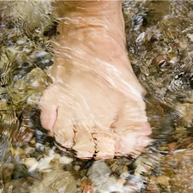 Ein Fuß im klaren Wasser, umgeben von kleinen Steinen. Die Wellen reflektieren sanft das Licht.