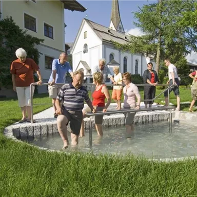 A group of people is standing around a small foot bath on a green meadow. In the background, there is a church and blue skies.