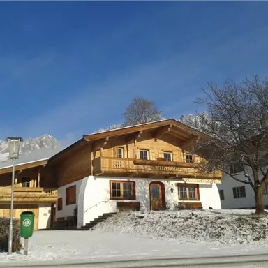 A typical alpine house with wooden cladding in a snowy environment. In the background, the mountains and a clear sky can be seen.