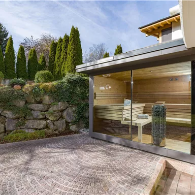 Eine moderne Sauna mit großen Glasfenstern, umgeben von einer gepflegten Gartenlandschaft. Im Hintergrund sind hohe Pflanzen und ein schöner Himmel sichtbar.