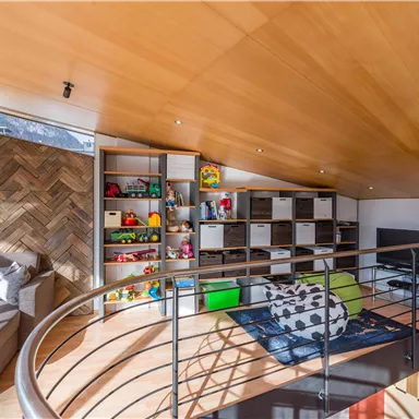 Ein moderner Raum im Dachgeschoss mit einem gemütlichen Sofa und einem offenen Regal voller Spielzeuge. Der Raum hat helles Holz und viel Tageslicht.