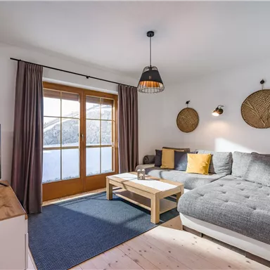 A cozy living room with a gray sofa and a wooden slatted table.
Large windows provide a view of the surroundings.