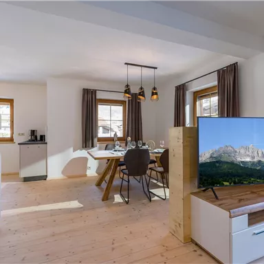 A modern living room with a large television, a dining table, and stylish chairs. Large windows bring plenty of daylight into the room.
