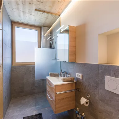 A modern bathroom with wood and gray tones. There is a sink, a mirror, and a window front that allows plenty of daylight in.