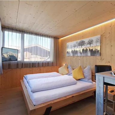 A modern bedroom with wood veneer, large windows, and bright daylight. Two single beds with yellow pillows and a writing area create an inviting atmosphere.