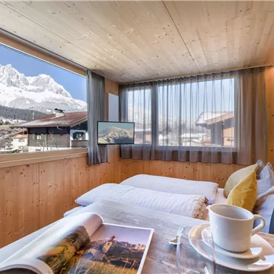 A cozy room with wooden walls and large windows. In the background, majestic mountains can be seen.