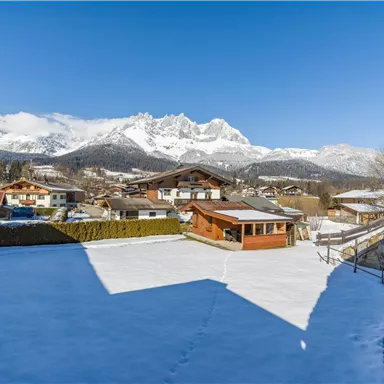 A snowy landscape with a view of the mountains. In the foreground, there are houses and a large lawn.