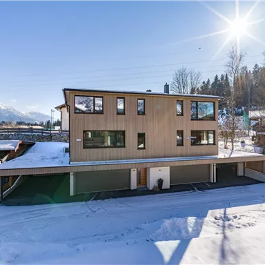A modern house in the snow with plenty of natural light. In the background, trees and mountains are visible.