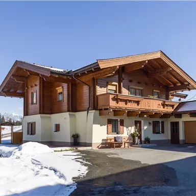 A modern wooden house in alpine style with a snow-covered front yard. The sky is clear and blue.