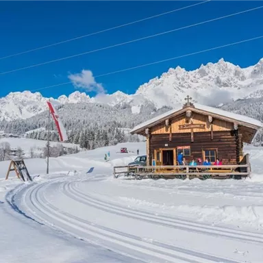 A cozy mountain cabin in the snow with impressive, snow-covered mountains in the background. The sky is clear and blue, creating a picturesque winter landscape.