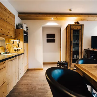 A modern kitchen with wooden cabinets and a dining table. In the background, a television and a shelf are visible.