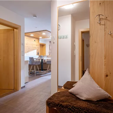 A modern hallway with wooden furniture and a cozy seating area. In the background, a dining area and a well-lit room are visible.