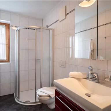 A modern bathroom with a shower cabin, sink, and a toilet. Light tiles and large windows create a friendly atmosphere.