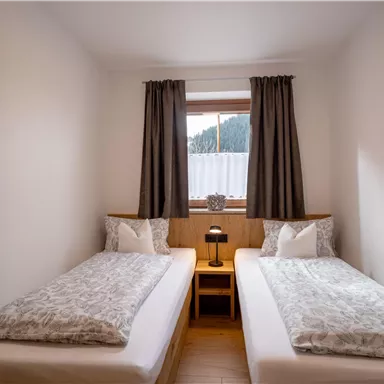 A bright bedroom with two single beds and a small bedside table. In front of the window, there are blackout curtains and a beautiful view of the mountains.