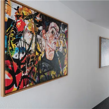 Ein farbenfrohes Kunstwerk hängt an einer weißen Wand im Flur. Im Hintergrund sind weitere Bilder und ein Fenster sichtbar.