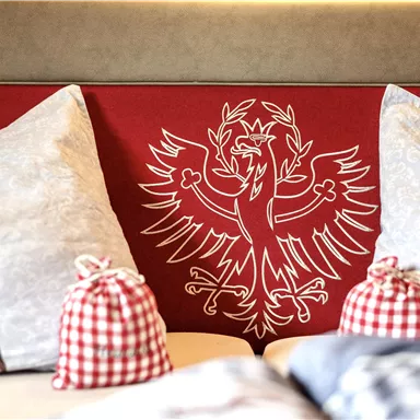 Ein gemütliches Zimmer mit dekorativen Kissen und einem roten Wanddesign. In der Mitte ist ein stilisiertes Wappen zu sehen.