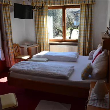 Ein gemütliches Schlafzimmer mit einem großen Bett und Fenster mit blickdichten Vorhängen. Der Raum hat einen warmen Farbton und enthält einen Stuhl sowie einen Tisch.