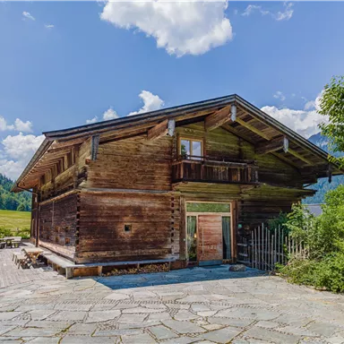 A traditional wooden house with a balcony and a large terrace. Surrounded by green meadows and trees under a clear blue sky.