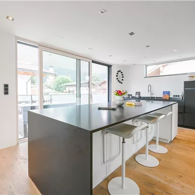 Moderne Küche mit einem großen, dunklen Kücheninsel und weißen Barhockern. Große Fenster sorgen für viel Tageslicht und Ausblick auf die Umgebung.